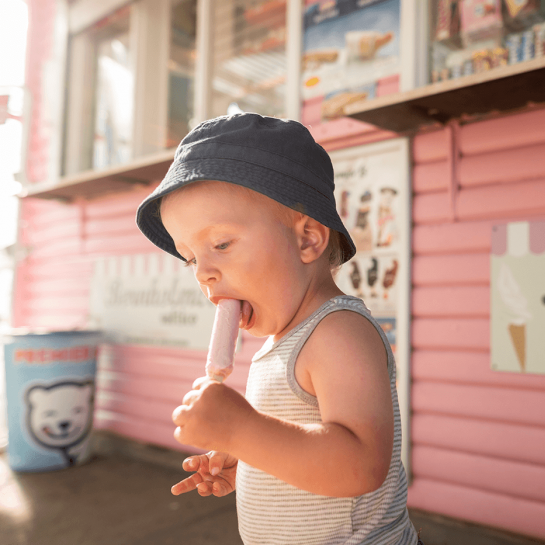 Lille barn med bøllehat spiser lyserød pindis ved kiosk