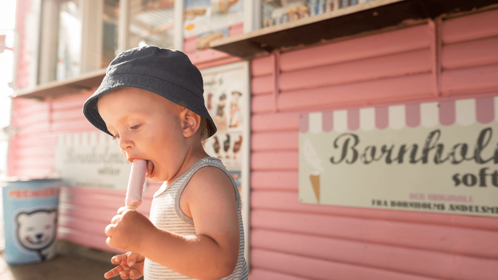 Lille barn med bøllehat spiser lyserød pindis ved kiosk